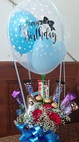 Blue Balloons stuffed inside a clear Printed with happy birthday balloon tied to a basket with 3 Ferrero rocher chocolates 3 Snickers 3 dairy milk and 3 red roses with Blue Ribbons