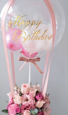 Pink small balloons inside a transparent hot air balloon printed happy birthday with pink roses and pink flowers basket