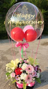Happy anniversary printed hot air balloon with small balloons inside tied to a basket with red gerberas white lily and white roses